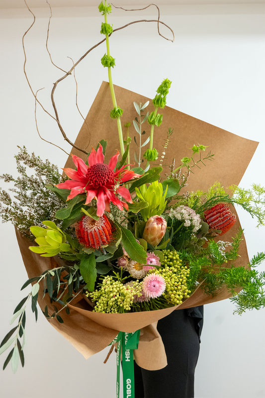 A vibrant mixed floral bouquet wrapped in brown kraft paper, featuring a prominent pink protea, red and yellow proteas, green eucalyptus, and other green foliage, hand crafted by GoBush Florist new farm, Brisbane.