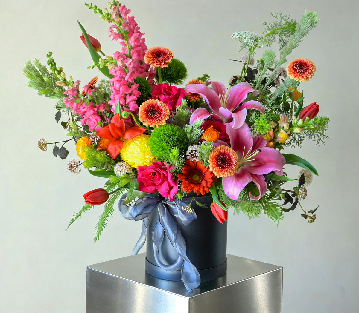 A vibrant, multicolored floral arrangement featuring pink lilies, pink snapdragons, orange gerbera daisies, red tulips, yellow lilies, and green foliage, presented in a black cylindrical box tied with a blue satin ribbon and displayed on a metallic silver pedestal against a neutral light gray background. designed by gobush florist brisbane