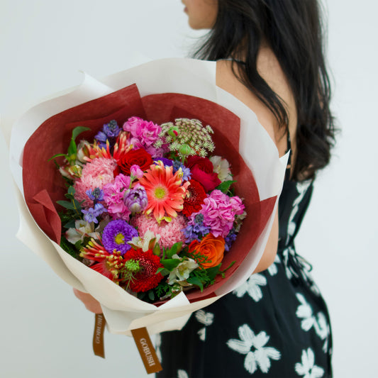 A vibrant, multicolored flower bouquet wrapped in white paper with a red interior liner. The bouquet features a variety of colorful flowers including pink, red, orange, purple, and green. designed by GoBush Florist Brisbane, in West end, Brisbane.