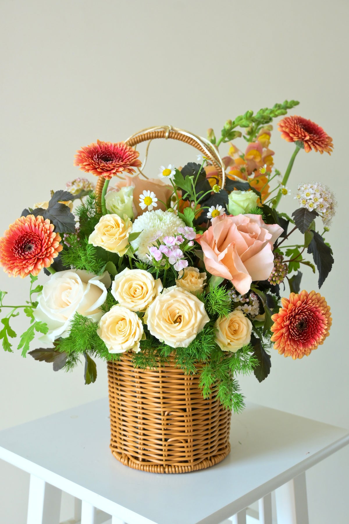 A wicker flower basket filled with assorted flowers, including peach roses, cream roses, orange gerbera daisies, and green foliage, displayed on a white table against a light beige wall.