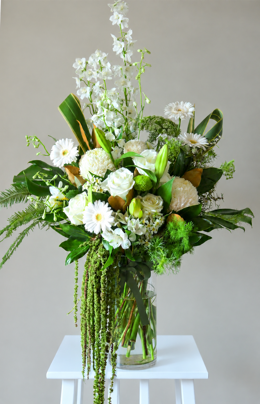 A large, lush white and green floral arrangement in a clear glass vase, displayed on a small white stool against a neutral light gray background.
