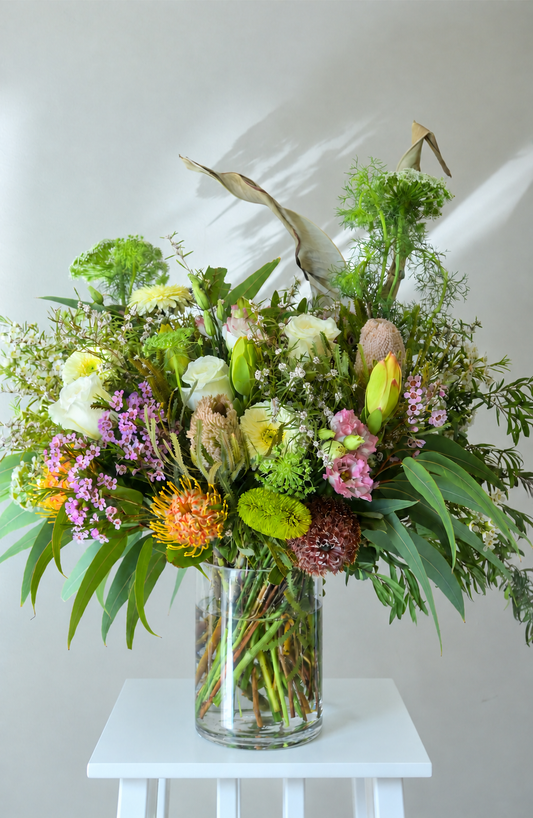 A large, vibrant wildflower and floral arrangement in a clear glass vase, displayed on a small white stool against a light gray wall with soft shadows.