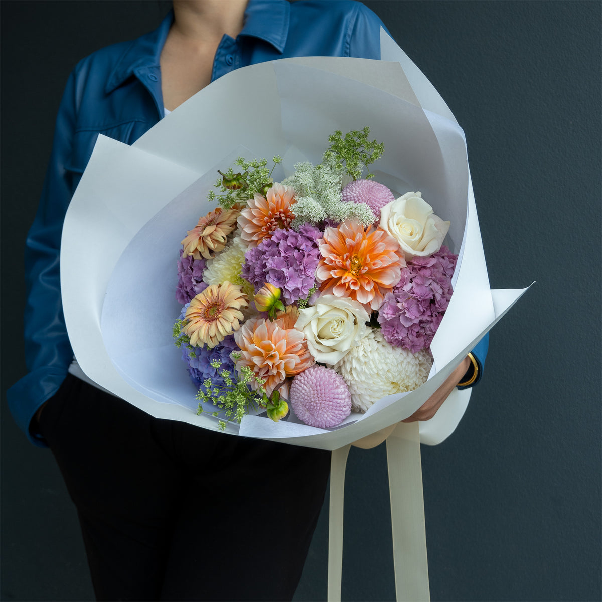 a large bouquet of mixed flowers wrapped in white paper. The bouquet contains orange, purple, white, and peach flowers with green foliage. Designed by GoBush Floral Studio, in west end, Brisbane.