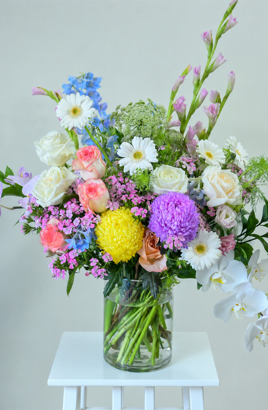 A vibrant mixed flower bouquet in a clear glass jar vase, displayed on a small white stool against a light gray background. The bouquet features a variety of colorful flowers including white daisies, pink and peach roses, blue delphiniums, yellow pompons, purple globes, and white orchids.