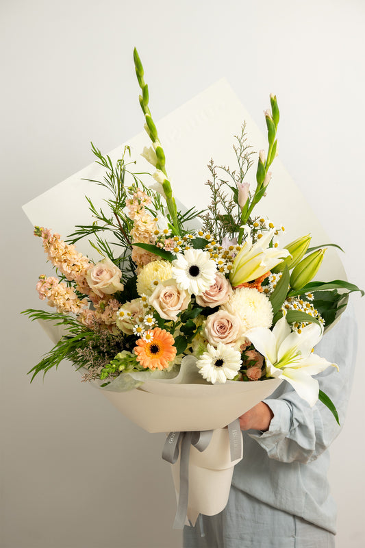 A large, beautifully arranged floral bouquet held by a person wearing a light blue shirt. The bouquet features a mix of white lilies, pale pink roses, orange daisies, and green foliage, wrapped in white paper with a gray ribbon.